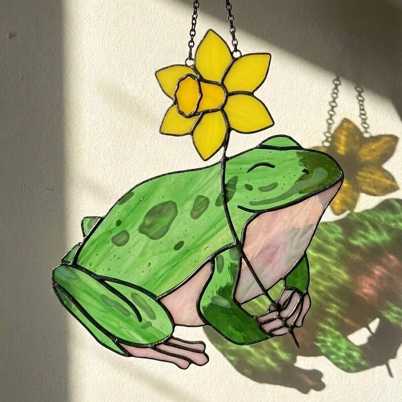May include: Stained glass frog suncatcher with a yellow daffodil flower. The frog is green with pink accents and a smiling face. The daffodil is yellow with a detailed center. The suncatcher hangs from a chain.