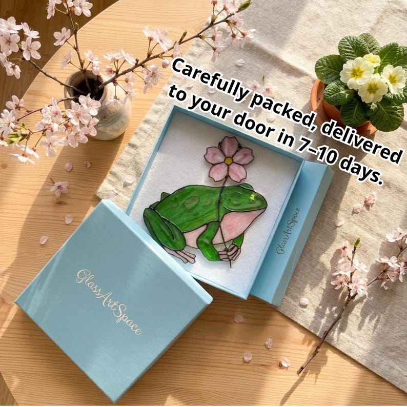May include: A stained glass frog holding a flower is displayed in a light blue box. The box is open, revealing the artwork on a white background. The text "Glass Art Space" is on the box. Cherry blossoms and a potted plant are in the background.