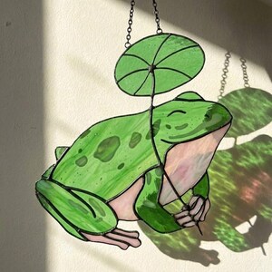 May include: A stained glass frog with a green lily pad umbrella. The frog is a vibrant green with darker green spots and pink accents. It is suspended by a chain, creating a whimsical decorative piece.