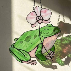 May include: Stained glass frog holding a pink and white orchid. The frog is green with darker green spots and pink accents. The orchid has pink and white petals. The piece is hanging from a chain.