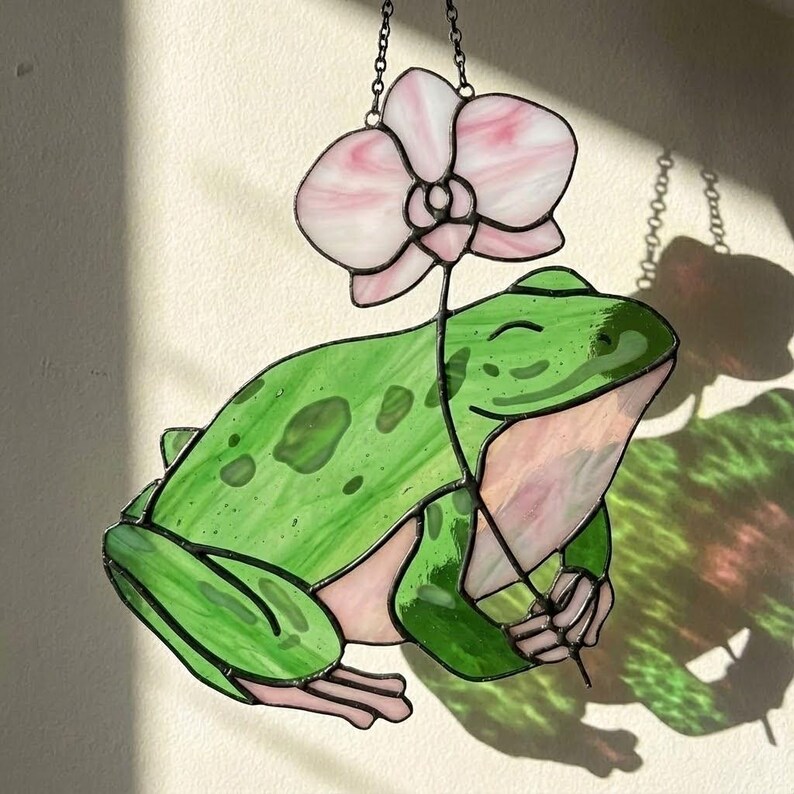 May include: Stained glass frog holding a pink and white orchid. The frog is green with darker green spots and pink accents. The orchid has pink and white petals. The piece is hanging from a chain.