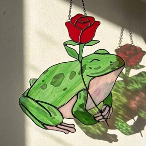 May include: Stained glass frog with a red rose. The frog is green with darker green spots and pink accents. The rose is a vibrant red. The piece is designed to hang, with a chain at the top.