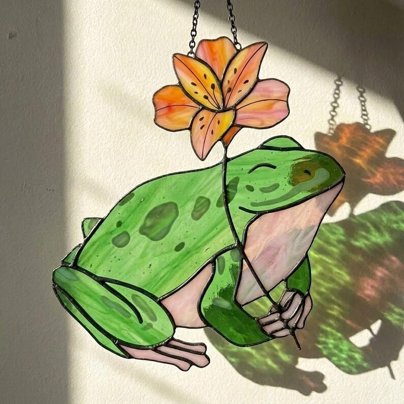 May include: Stained glass frog holding a flower. The frog is green with darker green spots and a pink belly. The flower is orange and pink with black details. The piece is hanging from a chain.
