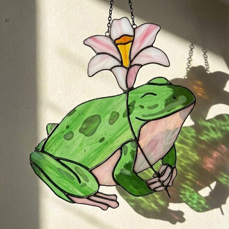 May include: Stained glass frog with a pink and white flower above its head. The frog is green with darker green spots and pink accents. The flower has pink petals and a yellow center. A chain is attached for hanging.
