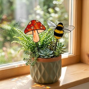 May include: A small potted succulent garden with decorative glass mushroom and bee stakes. The mushroom has a red and white spotted cap. The bee has yellow and black stripes and clear wings. The pot is green with a diamond pattern.