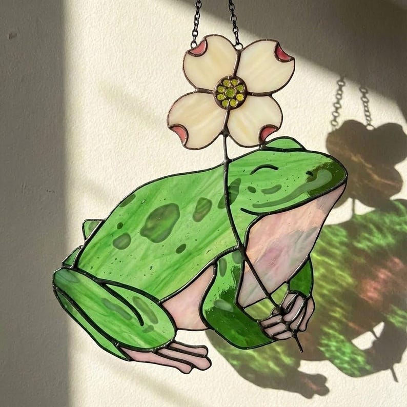 May include: Stained glass frog with a flower. The frog is green with darker green spots and pink accents. The flower is white with pink tips and a yellow center. The piece is hanging from a chain.