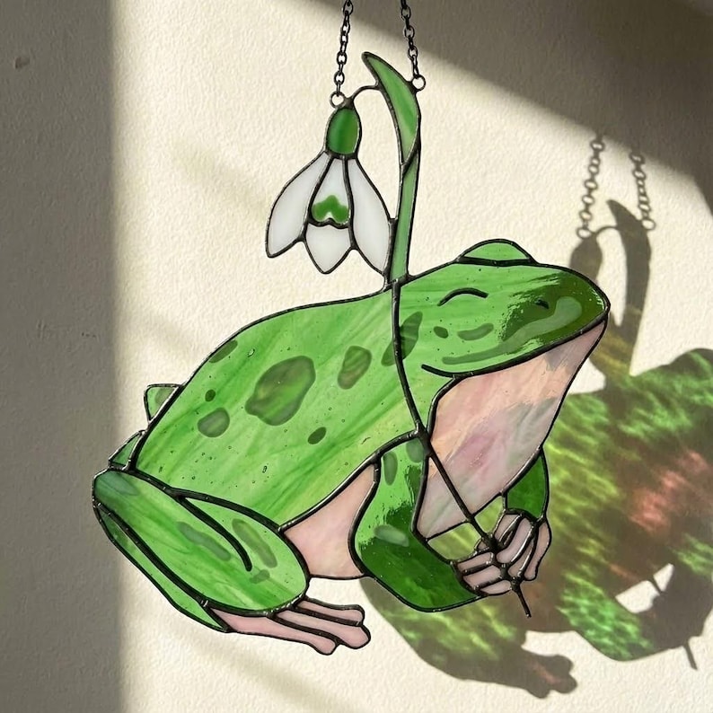 May include: A stained glass frog with a white flower, hanging from a chain. The frog is green with pink accents and a smiling face. The flower has white petals and a green stem. The piece is designed to hang in a window.