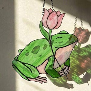May include: A stained glass frog with a pink tulip. The frog is green with darker green spots and pink accents. The tulip is pink and the piece hangs from a chain. The frog is smiling.