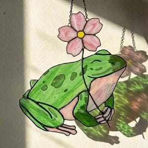 May include: Stained glass frog holding a pink flower. The frog is green with darker green spots and a light pink belly. The flower has pink petals and a yellow center. The piece is hanging from a chain.