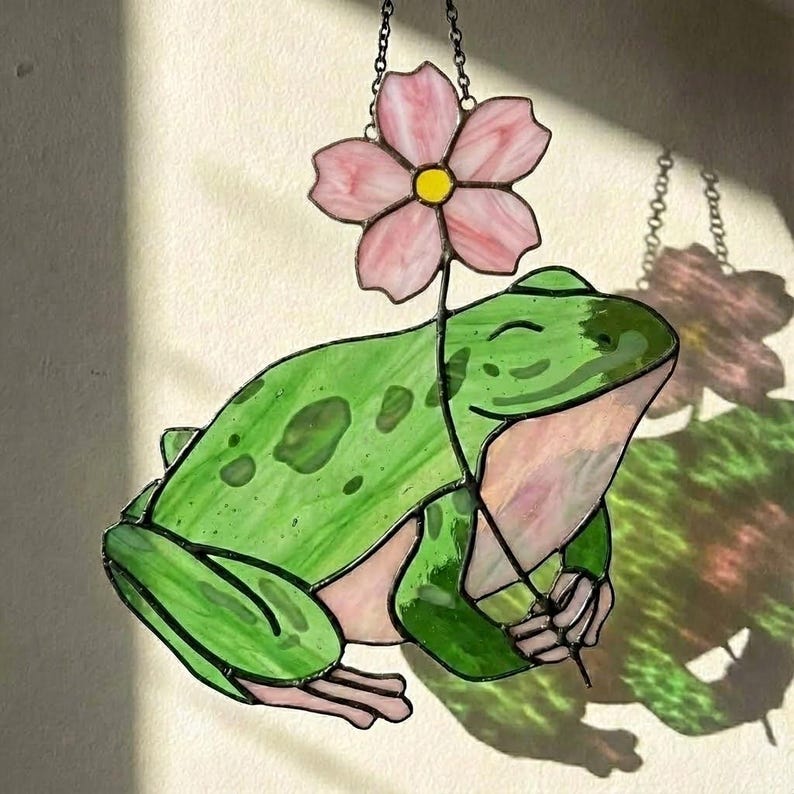 May include: Stained glass frog holding a pink flower. The frog is green with darker green spots and a light pink belly. The flower has pink petals and a yellow center. The piece is hanging from a chain.