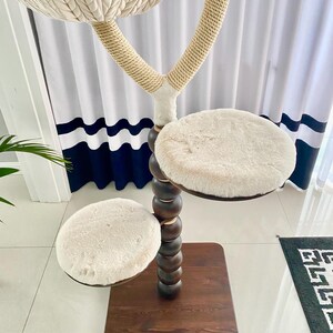 Pode incluir: Uma &aacute;rvore para gatos de madeira marrom com tr&ecirc;s plataformas cobertas de pele sint&eacute;tica branca. A plataforma superior &eacute; uma cama estilo rede. A &aacute;rvore &eacute; decorada com contas de madeira marrom.