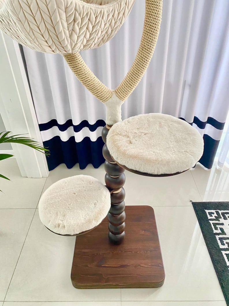 Pode incluir: Uma &aacute;rvore para gatos de madeira marrom com tr&ecirc;s plataformas cobertas de pele sint&eacute;tica branca. A plataforma superior &eacute; uma cama estilo rede. A &aacute;rvore &eacute; decorada com contas de madeira marrom.
