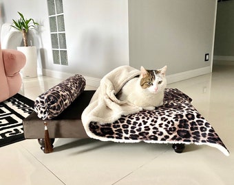 Può includere: Un letto per gatti marrone con un design a stampa leopardata. Il letto ha una coperta in pelliccia sintetica bianca e un cuscino marrone con un design a stampa leopardata. Un gatto bianco con macchie marroni è sdraiato sul letto.
