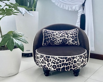 Può includere: Un letto rotondo per animali domestici con stampa leopardata marrone e bianca con un cuscino abbinato. Il letto ha una forma rotonda ed è rialzato su quattro gambe in legno.