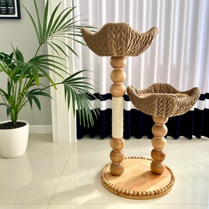 Pode incluir: Uma árvore para gatos de dois níveis com uma plataforma superior e inferior de tecido acolchoado bege. A árvore é feita de madeira e tem um poste coberto de corda no centro.