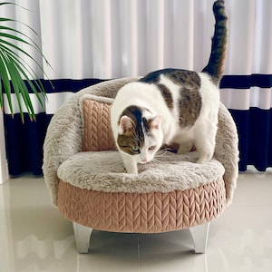 May include: A white and brown tabby cat standing on a plush, round pet bed with a brown and beige quilted exterior. The bed has a light gray base with silver legs.