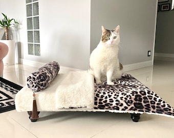 Può includere: Un gatto è seduto su un letto per animali con stampa leopardata con una coperta in finta pelliccia bianca e un cuscino a rullo abbinato. Il letto ha gambe in legno scuro e un nappa decorativa. Il gatto ha pelo bianco e marrone.