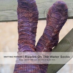 Pode incluir: Um par de meias de tricô roxas e marrons com um padrão ondulado. As meias estão penduradas em uma cerca de madeira. O texto "Knitting Pattern | Ripples On The Water Socks" e "Size: 36/37 (38/39) (40/41) // 3.00 mm" está impresso abaixo das meias.