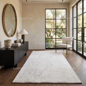 Peut inclure: Intérieur moderne avec un grand tapis rectangulaire clair à motif géométrique. La pièce comprend un meuble en bois foncé, un miroir rond, une lampe et une chaise. De grandes fenêtres offrent une vue sur l'extérieur.