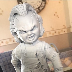May include: A gray figurine of a doll with stitched scars on its head, wearing overalls and holding a small weapon. The doll has a sinister grin and is standing on a circular base.