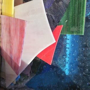 Op de afbeelding: Diverse stukken glas-in-lood in verschillende vormen en kleuren, waaronder geel, rood, wit, roze, groen en blauw. Het glas zit in een kartonnen doos, klaar voor verzending.