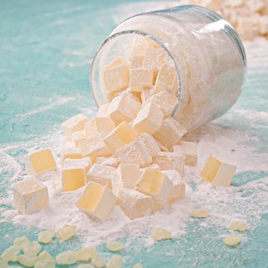Könnte beinhalten: Ein umgestürztes Glasgefäß, aus dem zahlreiche quadratische, hellgelbe türkische Honig-Bonbons herausfallen, die mit Puderzucker bestäubt sind. Die Süßigkeiten sind auf einer hellblauen Oberfläche verstreut.