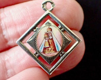 Peut inclure: Pendentif en argent avec un cadre en forme de losange. À l'intérieur, un cadre carré plus petit contient une image religieuse de deux figures couronnées. L'image est dans les tons de rouge, d'or et de bleu. Le pendentif a une petite boucle en haut.