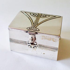 May include: A small, rectangular silver box with a hinged lid. The lid features a stylized design. The box has horizontal lines and a decorative clasp with a cluster of small, round details. The box is likely used for jewelry or small keepsakes.