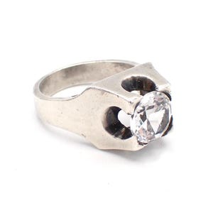 Puede incluir: Un anillo de tono plateado con una piedra redonda, transparente y facetada, engastada en una banda de metal gruesa y sólida. El engaste presenta un diseño arquitectónico distintivo con aberturas laterales que enmarcan la piedra central.