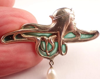 May include: A vintage Art Nouveau style brooch featuring a woman's profile in gold-tone metal with flowing hair and a light green enamel background. A teardrop-shaped pearl dangles from the bottom of the brooch.
