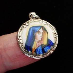 Puede incluir: Colgante redondo plateado con una imagen de esmalte de una mujer con velo azul y chal amarillo. El colgante tiene un borde decorativo y un pequeño lazo para una cadena. El fondo es negro.