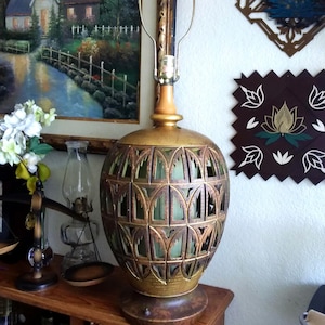 May include: A vintage table lamp with a gold-colored metal base and a decorative, openwork shade. The shade has an arched design with a green interior. The lamp is on a wooden table, with a painting and other decorative items in the background.