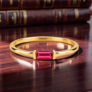 Può includere: Un sottile anello in oro con una singola pietra preziosa rosa a taglio baguette rettangolare incastonata orizzontalmente al centro. Il metallo ha una finitura gialla lucida e la pietra è di una tonalità rosa vibrante e satura.