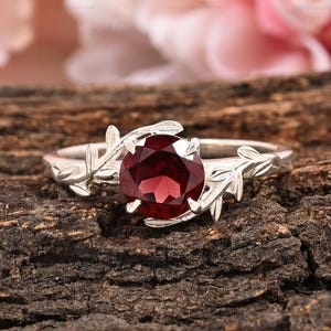 Pode incluir: Um anel de prata com uma grande pedra preciosa redonda e vermelha escura. O anel apresenta um design de folhas em ambos os lados da pedra preciosa. O fundo é uma imagem desfocada de flores cor-de-rosa e uma superfície de madeira castanha escura.