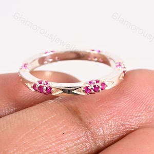 May include: A silver ring with a band of small, bright pink gemstones. The ring has a repeating pattern of triangular cutouts and clusters of gemstones. The ring is held in a person's hand.