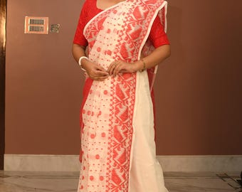 May include: A woman wearing a red blouse and a white and red patterned saree. The saree features intricate floral and geometric designs. The outfit is completed with a red necklace and bracelets. The background is a plain brown wall.