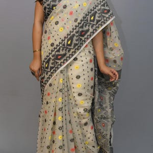 May include: A white saree with black, red, and yellow floral patterns. The saree has a black border with geometric designs.