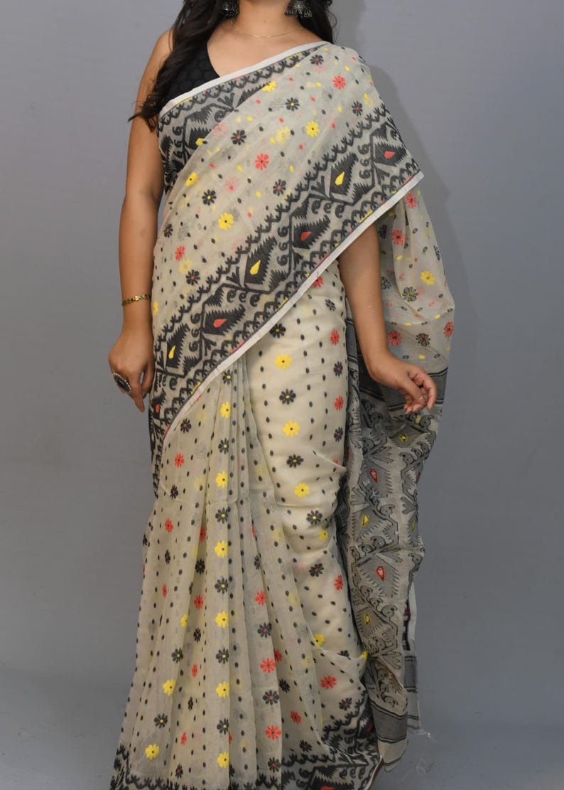 May include: A white saree with black, red, and yellow floral patterns. The saree has a black border with geometric designs.