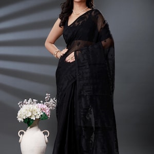 Może przedstawiać: Kobieta w czarnym sari z przezroczystą nakładką. Sari ma subtelny wzór i układa się elegancko. Ma na sobie złotą biżuterię i perłowy naszyjnik. Biały wazon z kwiatami w tle.