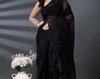 May include: A woman wearing a black saree with a sheer overlay. The saree has a subtle pattern and drapes elegantly. She is wearing gold jewelry and a pearl necklace. A white vase with flowers is in the background.