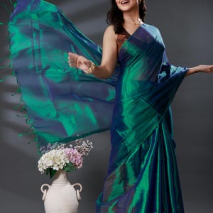 May include: A flowing, iridescent green and blue saree. The fabric drapes elegantly, with a shimmering effect. The saree has a fringe detail at the edge. The overall look is enhanced by the play of light on the fabric.
