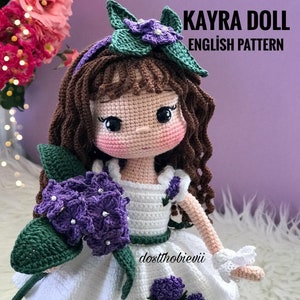 May include: Crochet doll pattern for a girl doll with brown hair, a white dress, and purple flowers. The pattern is called "Kayra Doll English Pattern".