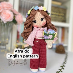May include: A crocheted doll with brown hair, a pink shirt, red trousers, and a flower crown. The doll is holding a bouquet of flowers. The doll is called "Afra doll" and the image includes the text "English pattern".