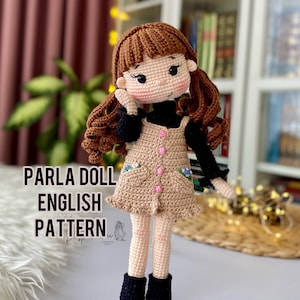 May include: Crocheted doll with brown hair, a black shirt, a beige dress with pink buttons, and black boots. The doll is standing on a table with a woven mat. The text "PARLA DOLL ENGLISH PATTERN" is visible in the image.