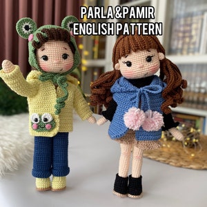 May include: Two crocheted dolls, one wearing a green frog hat and yellow jacket, the other wearing a blue vest with pink pom-poms. The dolls are standing on a white surface. PARLA & PAMIR ENGLISH PATTERN.