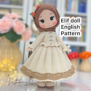 Può includere: Una bambola all'uncinetto fatta a mano con hijab marrone e vestito color crema. La bambola ha un'espressione dolce con guance rosee e grandi occhi. Il testo "Elif doll English Pattern" è visibile.