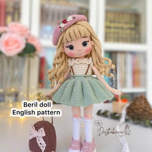 Peut inclure: Une poupée en crochet aux cheveux blonds, portant un béret rose, une robe verte et des chaussettes blanches. La poupée tient un sac à dos brun avec un nœud rose. La poupée est debout sur une surface blanche. Le texte "Beryl doll English pattern" est visible sur l'image.