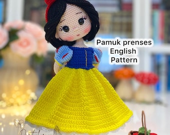May include: A handmade crochet doll of Snow White, featuring a yellow dress, blue bodice, and red bow. The doll has black hair and red shoes. Text on the image reads "Pamuk prenses English Pattern". A small basket of crochet strawberries is also present.