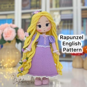 May include: A handmade crochet doll of Rapunzel with long yellow hair, wearing a purple dress. The doll has a sweet expression and is adorned with small floral accents. The text "Rapunzel English Pattern" is visible.