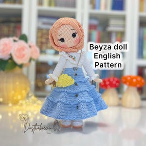 May include: A crocheted doll with a peach head covering and rosy cheeks. The doll is dressed in a white top, a blue skirt, and a yellow purse. The text "Beyza doll English Pattern" is displayed.